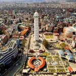 An aerial drone perspective looking down at the newly reconstructed white Dharahara Tower and its modern landscaped public park grounds in downtown Kathmandu, Nepal.