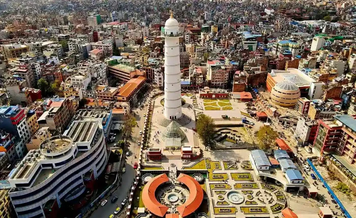 An aerial drone perspective looking down at the newly reconstructed white Dharahara Tower and its modern landscaped public park grounds in downtown Kathmandu, Nepal.