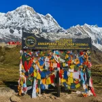 The official black welcome signboard at Annapurna Base Camp decorated with colorful Buddhist prayer flags against a backdrop of snow mountains.