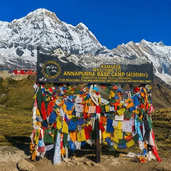 The official black welcome signboard at Annapurna Base Camp decorated with colorful Buddhist prayer flags against a backdrop of snow mountains.