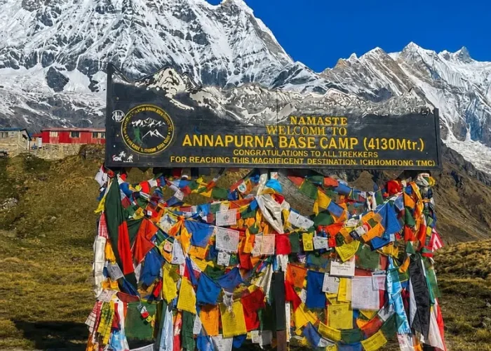 The official black welcome signboard at Annapurna Base Camp decorated with colorful Buddhist prayer flags against a backdrop of snow mountains.
