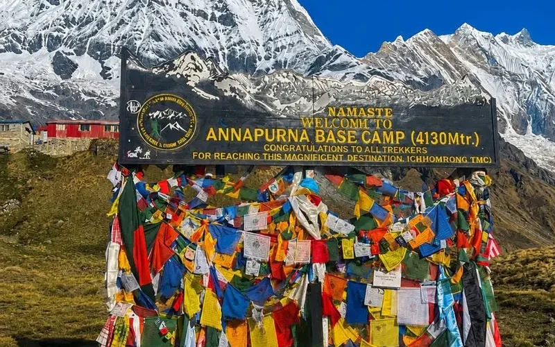 The official black welcome signboard at Annapurna Base Camp decorated with colorful Buddhist prayer flags against a backdrop of snow mountains.