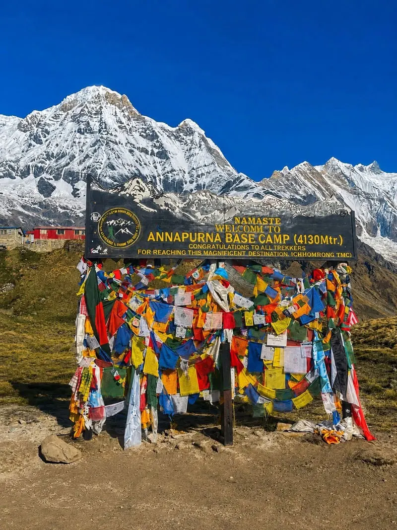 The official black welcome signboard at Annapurna Base Camp decorated with colorful Buddhist prayer flags against a backdrop of snow mountains.