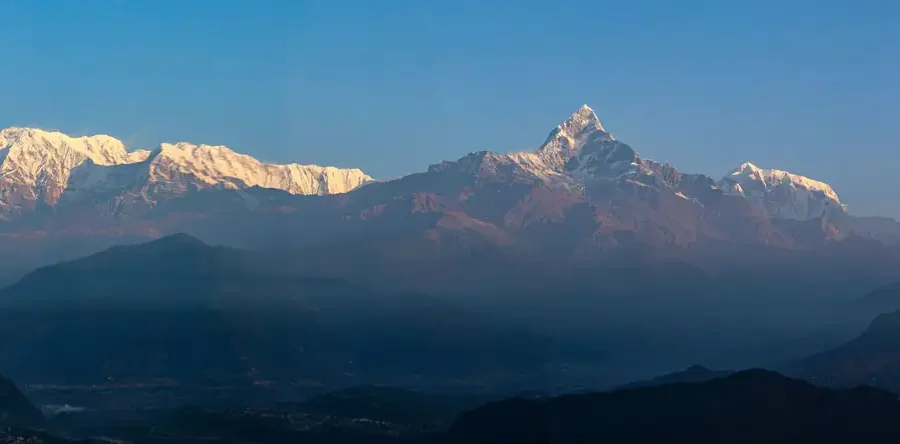 A wide panoramic landscape of the snow-capped Annapurna mountain range rising above the hazy blue layers of the Pokhara valley hills.