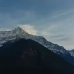 A wide landscape view of the massive Annapurna mountain range and the sharp peak of Mount Machapuchare rising above dark forested hills.