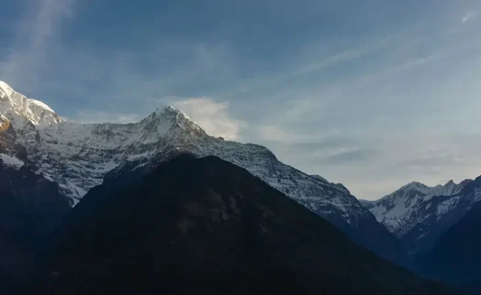 A wide landscape view of the massive Annapurna mountain range and the sharp peak of Mount Machapuchare rising above dark forested hills.