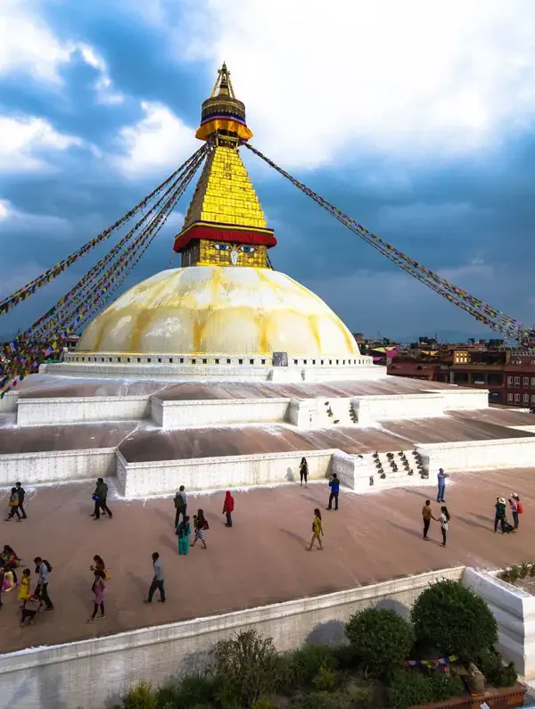Tourists and Buddhist pilgrims walking along the upper tiers of the massive white dome of Boudhanath Stupa under dramatic clouds.