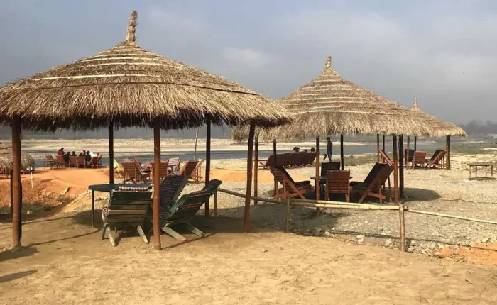 Traditional thatched-roof umbrellas and beach chairs at a riverside resort lounge area along the Rapti River banks in Sauraha.