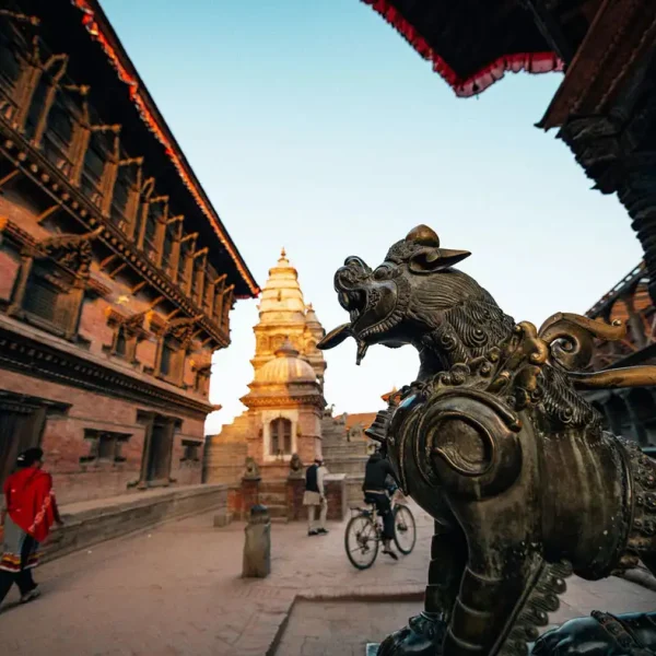 A close-up of a brass guardian lion statue with the historic 55 Window Palace and National Art Gallery in the background, Bhaktapur.