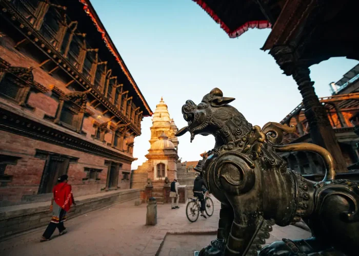 A close-up of a brass guardian lion statue with the historic 55 Window Palace and National Art Gallery in the background, Bhaktapur.