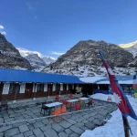 A mountain teahouse lodge with a blue roof at Machapuchare Base Camp, featuring a fluttering Nepal flag against a backdrop of snowy valley walls.