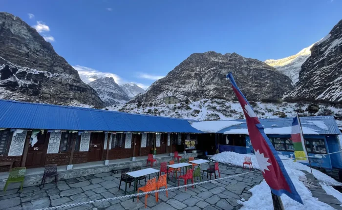 A mountain teahouse lodge with a blue roof at Machapuchare Base Camp, featuring a fluttering Nepal flag against a backdrop of snowy valley walls.