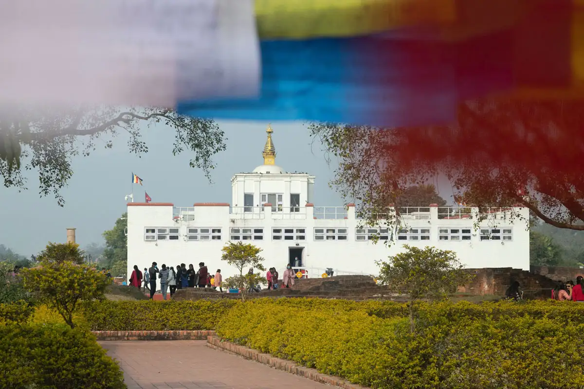 Tourists walking along a path toward the white Maya Devi Temple, the birthplace of Lord Buddha, framed by prayer flags.