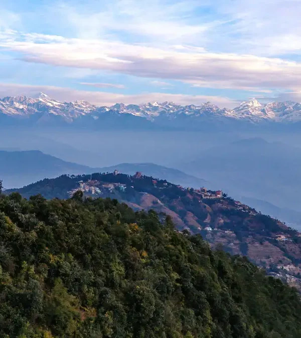 Panoramic view from a Nagarkot hilltop featuring the Nepal national flag waving on a ridge overlooking rolling hills and a massive snow-covered mountain range.