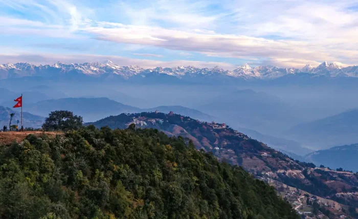 Panoramic view from a Nagarkot hilltop featuring the Nepal national flag waving on a ridge overlooking rolling hills and a massive snow-covered mountain range.