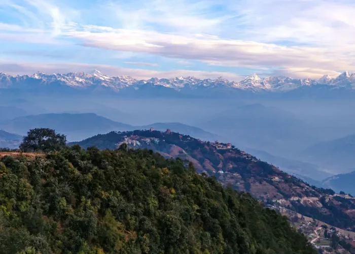 Panoramic view from a Nagarkot hilltop featuring the Nepal national flag waving on a ridge overlooking rolling hills and a massive snow-covered mountain range.