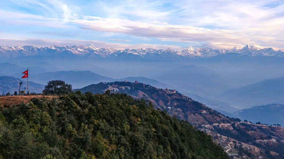 Panoramic view from a Nagarkot hilltop featuring the Nepal national flag waving on a ridge overlooking rolling hills and a massive snow-covered mountain range.