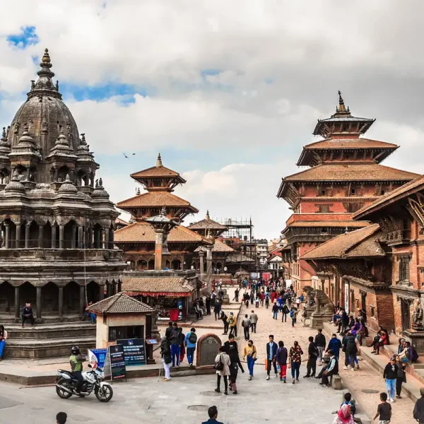 A lively daytime view of Patan Durbar Square showing the multi-tiered stone Krishna Mandir temple with visitors walking around.
