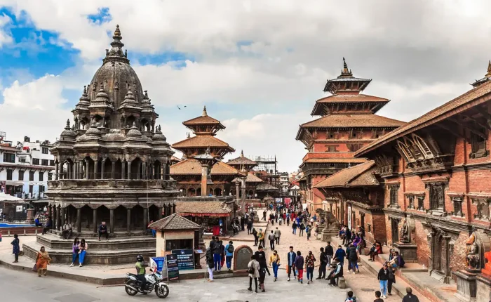 A lively daytime view of Patan Durbar Square showing the multi-tiered stone Krishna Mandir temple with visitors walking around.