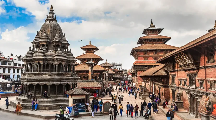 A lively daytime view of Patan Durbar Square showing the multi-tiered stone Krishna Mandir temple with visitors walking around.