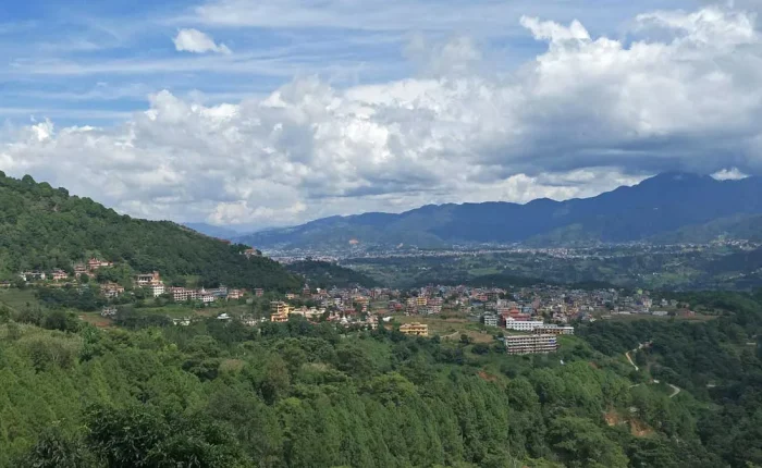 A sweeping panoramic landscape view of the hills, monastic towns, and valley settlements of Pharping under a dramatic cloudy blue sky.