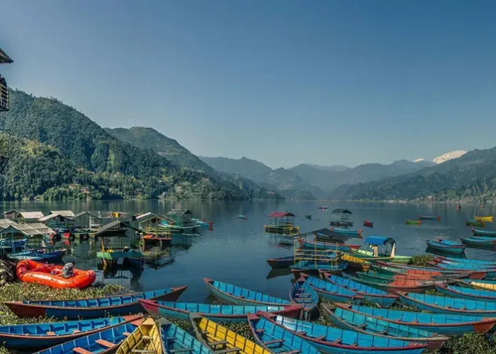 A wide panoramic landscape showing a lakeside boat station tower and dozens of colorful wooden canoes parked along Phewa Lake with mountains in the distance.