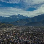 An aerial panoramic view of the sprawling Pokhara cityscape nestled beneath the massive peaks of the Annapurna mountain range.