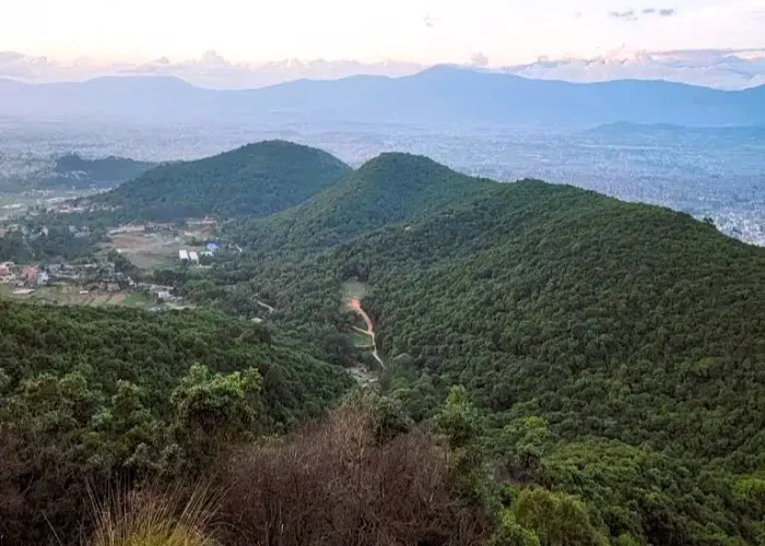 An expansive landscape view of lush green forested ridges and rolling hills stretching toward the Kathmandu valley horizon under a soft sunset sky.